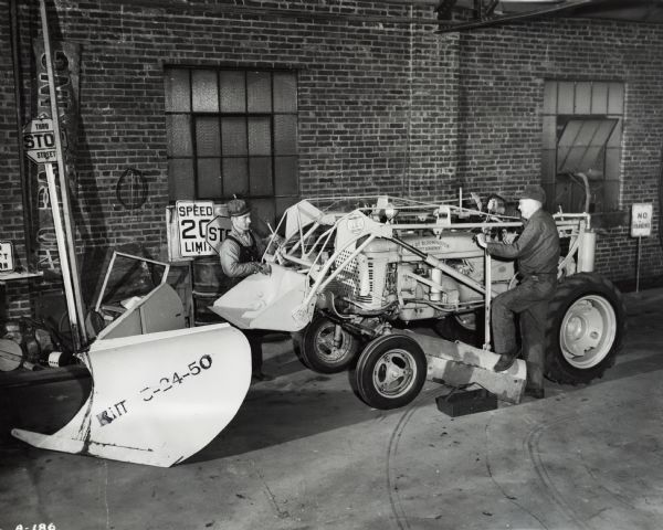 Two men are standing with an International A tractor owned by the City of Bloomington. The tractor has a grader and front loader attached. The men are preparing to attach a snow plow to the tractor. Street signs are leaning up against the wall. Original caption states: "All-around utility. Front end loader, dozer. Gathers leaves with grader (has board attached so as not to damage streets). Loader is used to move cinders from stockpile into trucks for spreading on streets in winter. Pix show street dept. mechanic Delmar Medlock and Joseph Fulford (sweeper driver) removing grader and loader to install snow plow. Only unit of its type owned by dept., which also has Galion grader."
