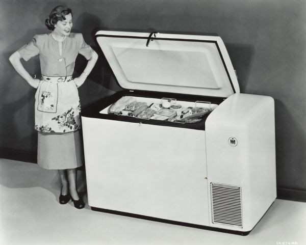 A woman in a dress, heels, apron, and pearls poses next to an open International Harvester freezer. The freezer, a unit with the lid on top, is full of food items.