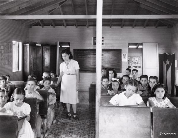 An interior view of "the village school" on an International Harvester sisal plantation in Cuba. A teacher stands while a classroom full of students sit at their desks. There are two Cuban flags at the back of the room.