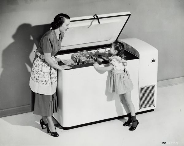 A woman in a dress, apron, heels, and earrings leaning over an open International Harvester freezer, smiling at a girl. The girl, in a dress, braided pigtails, and shiny black shoes, is standing on her toes, pointing to the back of the freezer.