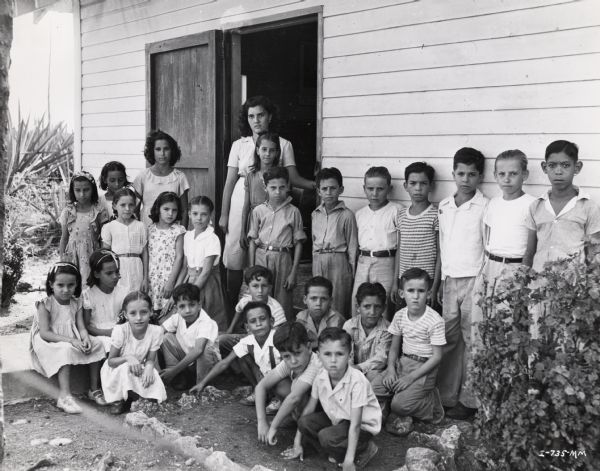 Students and teacher lined up outside a school building on an International Harvester sisal plantation in Cuba. Original caption reads: "School teacher and children lined up for their picture outside the village schoolhouse."
