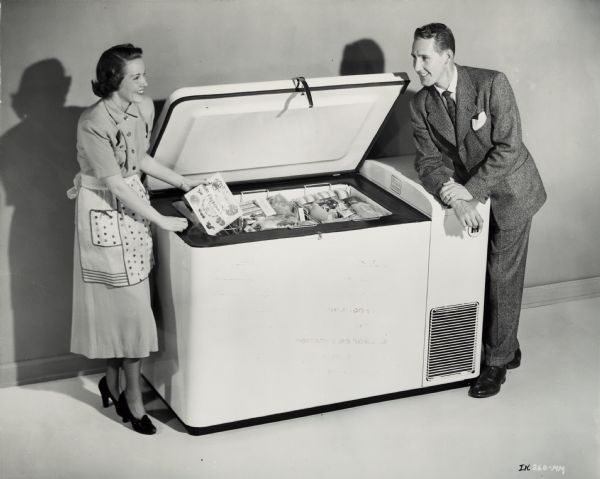 A woman in an apron, dress, and heels, standing on one side of an open International Harvester freezer with a food package in her hand. On the other side, a man is leaning on top of the freezer, dressed in a suit.  Both are smiling and looking towards each other.  Food packages are in the freezer.