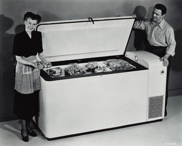 A woman is standing on the left in a dress, apron, and heels holding packaged food. On the right is a man standing on the other side of an open International Harvester freezer.