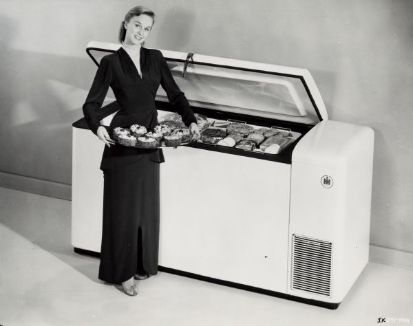A woman in a long dress, heels, earrings, and a double strand of pearls is holding of platter of food in front of an open International Harvester freezer.