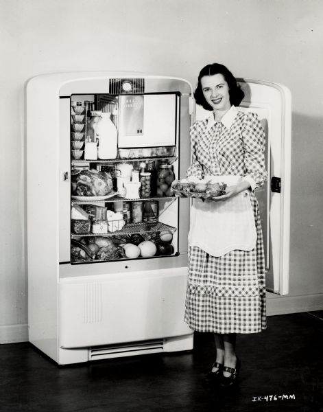 A woman in a checkered dress, apron, and heels is standing in front of an open International Harvester refrigerator.  The door is behind her, and she is holding a tray of food.  In the fridge is a variety of foods, including milk, jello cups, a dressed ham, fruits, vegetables, eggs, and canned items.