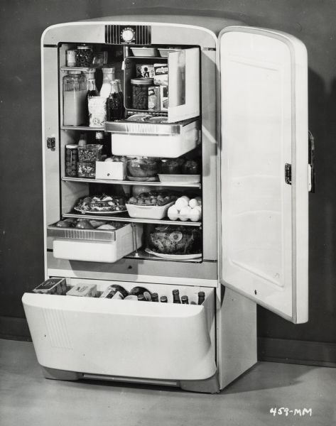 An International Harvester refrigerator with all doors open sits against a wall. The refrigerator has two drawers, a small freezer, and food. Bottles and cans are outside the refrigerator, including milk, juice, eggs, a dressed ham, stuffed peppers, and canned items.