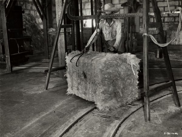 A man wearing a cowboy hat is using hooks on the end of a chain to move a bale of sisal fiber on an International Harvester plantation in Cuba.  A curved track is in the foreground, and another is in the background.