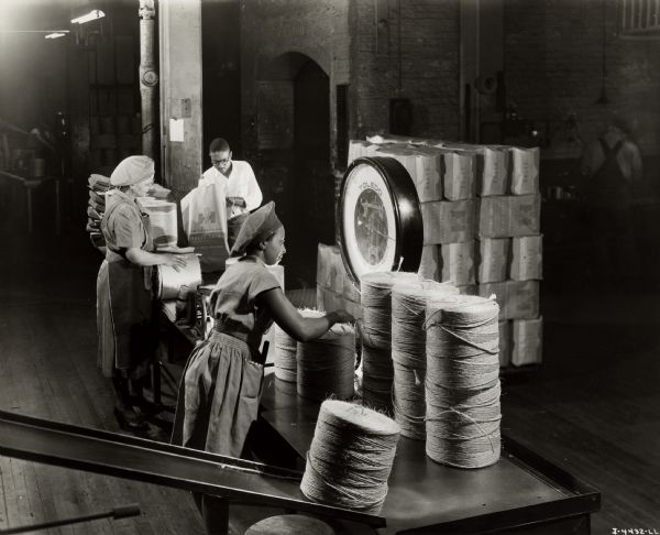 Male and female workers weighing bundles of twine on a scale, possibly at the McCormick Twine Mill in Chicago.