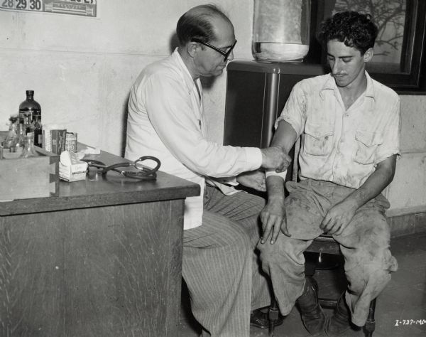 Doctor applying a bandage to the arm of a worker at an International Harvester sisal plantation in Cuba. On the left is a table with a stethoscope and medical supplies.