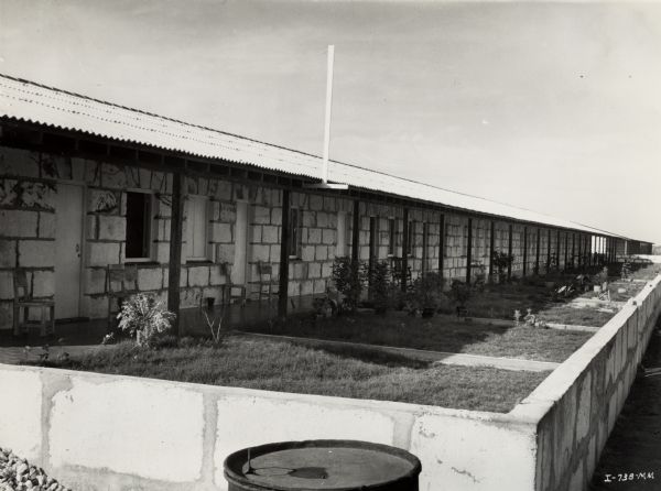 A long building containing workers quarters at an International Harvester sisal plantation in Cuba. The homes all share a long, covered porch. The lawns are separated by low boards, and end against a stone wall.