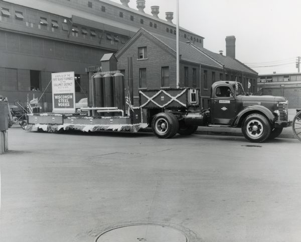 Parade float with a replica of the "first blast furnace in the Calumet district, built in 1875, the forerunner of Wisconsin Steel Works." The float is hitched to a 1949 model International truck. Factory buildings are in the background.