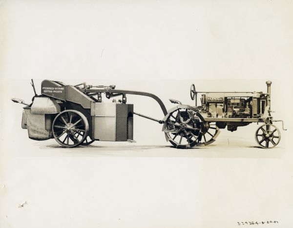 Studio shot of a Farmall Regular tractor and an experimental spindle type cotton picker. Original caption reads: "New McCormick-Deering Spindle Type Cotton Picker Pulled and Operated by General-Purpose Farmall Tractor. This machine is still in the semi-experimental stage  of development. It has been designed for use in the lowlands and other sections of the Old South where the entire crop cannot be picked at one time owing to long season and uneven ripening. The machine is equipped with two vertical picking cylinders, which carry a large number of spindles. These spindles work horizontally and are close enough together so that at least one of them will come in contact with every open boll on the plant. The mechanism of the picker is operated by power taken directly from the tractor engine by power take-off shaft."