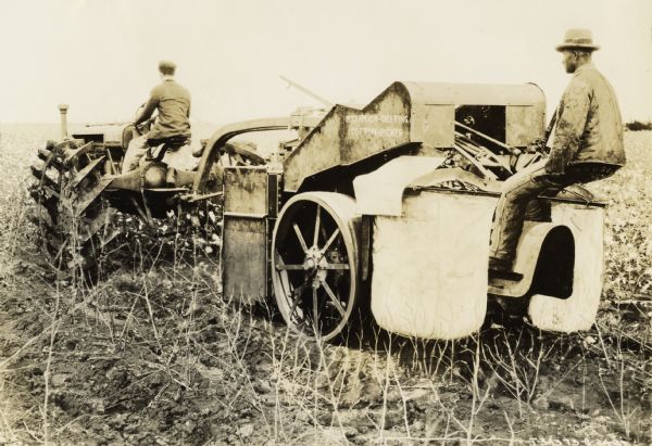 Two men in a cotton field with a Farmall Regular tractor and an experimental cotton picker. Original caption reads: "McCormick-Deering Cotton Picker and Farmall in Field of Cotton. As the picker is pulled along the cotton row, two large gathering shoes on the front of the machine pick up the spreading branches of the plants and place them in position for the picking spindles on the two cylinders. After passing through cleaning devices, the cotton is delivered into the two gathering bags as shown at rear of machine."