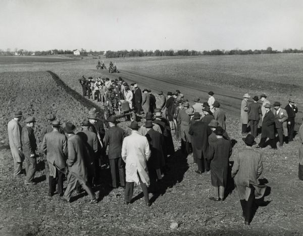 A group of spectators gathers around the edge of a field on the Hinsdale experimental farm to view a tractor in use.