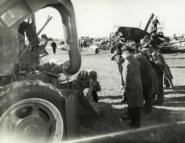 A group looks on as a man removes what appears to be a burlap bag from a piece of agricultural equipment on International Harvester's Hinsdale experimental farm.