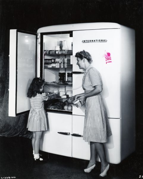 A woman and girl standing by an International Harvester upright refrigerator loading food onto the shelves. A curtain is in the background.