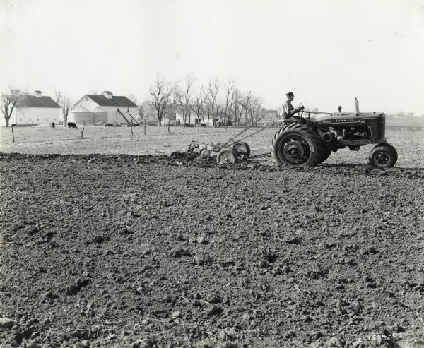 View from right of Clem J. Reynolds using a Farmall MD tractor and a No. 8 Little Genius plow to work in a field. Farm buildings and livestock are in the background.