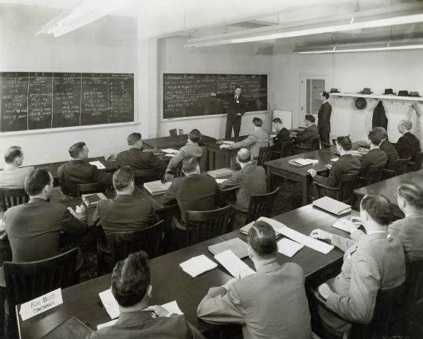 Rear view of group of employees sitting at tables to watch as an instructor demonstrates a lesson on a chalkboard at International Harvester's Education and Training Center.
