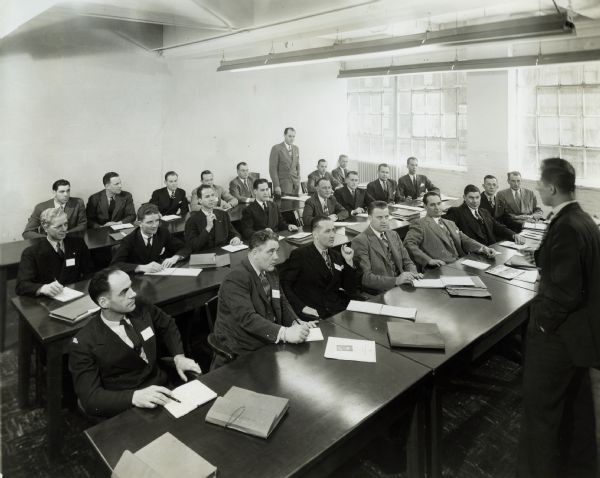 A group of male employees sitting at tables in International Harvester's education and training center while an instructor is speaking to them.