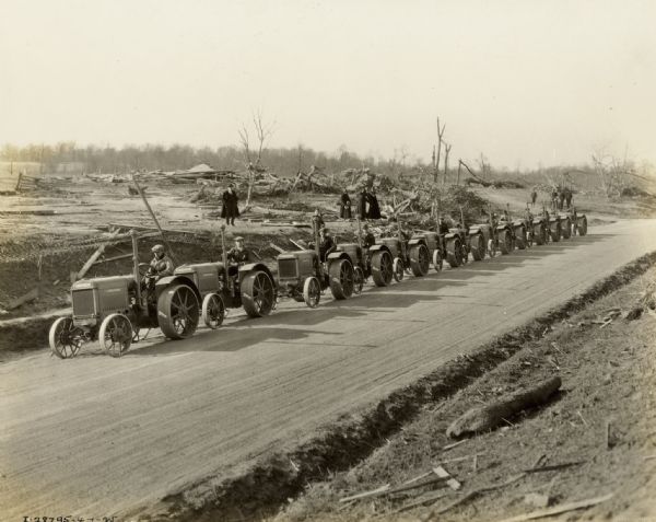 Men driving thirteen McCormick-Deering relief tractors down a dirt road for delivery to the victims of the "tri-state tornado." There are people watching from the left side of the road, and damaged trees and debris can be seen. The tractors appear to be 10-20s.
