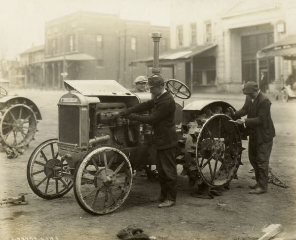 Three men working on a McCormick-Deering 10-20 relief tractor. They are working in the street, and storefronts are behind them.