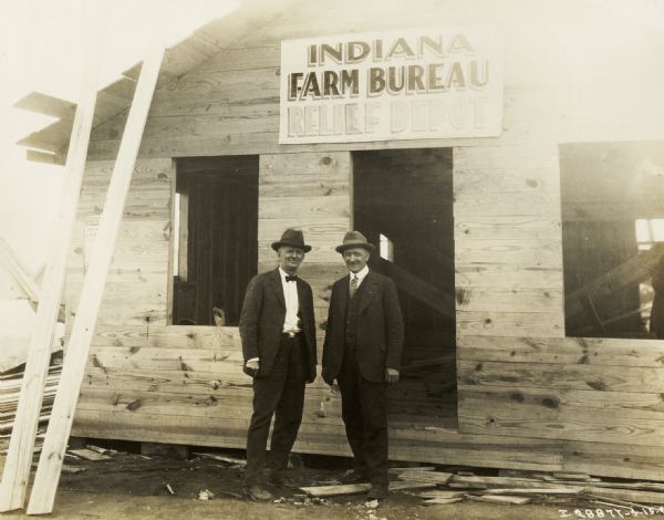 Two men standing outside the Indiana Farm Bureau relief depot. The building appears to be under construction, with no doors or windows in place, and lumber leaning against the roof.