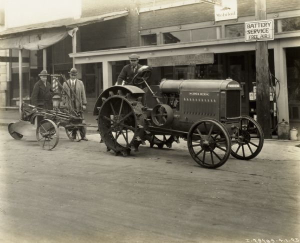Three men posing around a McCormick-Deering tractor in front of a Mobiloil station on a road. The tractor appears to be a 10-20.