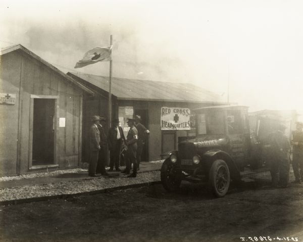Men are standing under a flagpole at the entrance of the Red Cross Headquarters. An International Red Baby truck is parked nearby.