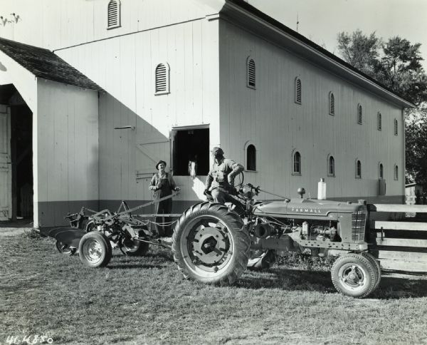 Charles Miller sitting on a McCormick-Deering Farmall H tractor and looking over his shoulder towards his son who is standing near a barn. A horse is looking out a window of the barn.