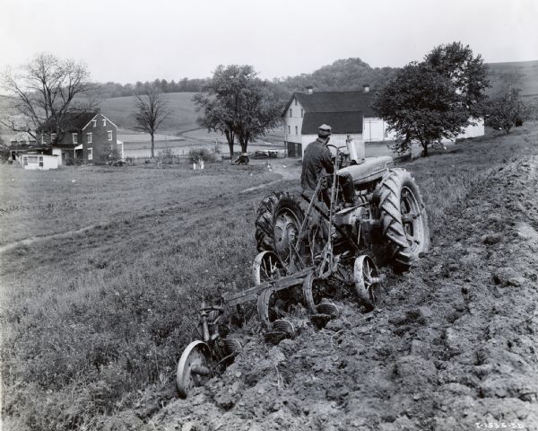 Rear view of a man using a McCormick-Deering Farmall M tractor with a Number 8 bottom plow to do field work on a hillside. Farm buildings are in the background.