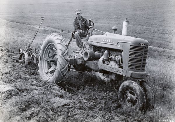 A man uses a McCormick-Deering Farmall M tractor with a Number 8 bottom plow to work in a field.