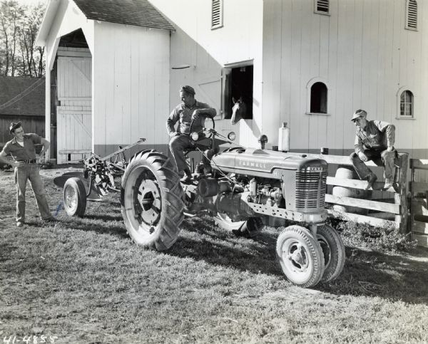 Charles Miller sitting on a McCormick-Deering Farmall H tractor while Gus Setser is standing behind him and James Miller is sitting on a fence. A horse is looking out the window of a barn in the background.