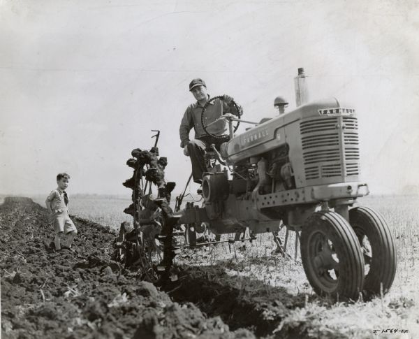 John Hanna uses a Farmall H with lug wheels and a Little Genius 2-14 bottom plow to work in a field while a boy looks on from the left.