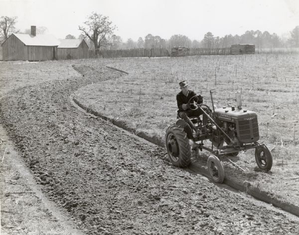 Slightly elevated view of Mason Rorbett using a McCormick-Deering Farmall A tractor with a 92 slab moldboard plow to build a terrace on his farm. In the background are farm buildings and a fence.