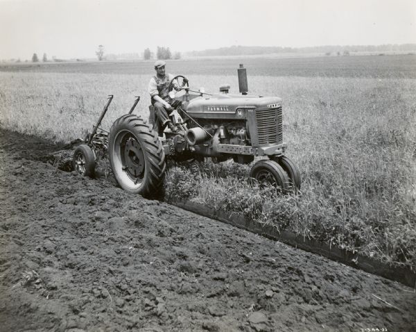 Three-quarter view from front right of a farmer using a McCormick-Deering Farmall M tractor with a Little Genius plow to work in a field on the Bigler farm.