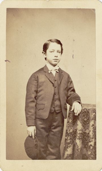 Three-quarter length studio portrait of a boy wearing a suit jacket, necktie, vest, and trousers. He is holding a hat while posing next to a table. Possibly Leander Hamilton McCormick (1859-1934).