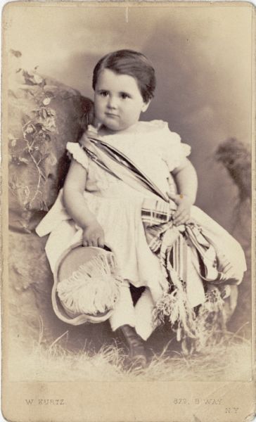 Carte-de-visite portrait of Harold Fowler McCormick as a young boy, (1872-1941) posing holding a hat with a large feather, and wearing a sash over his shoulder which is tied at his waist.
