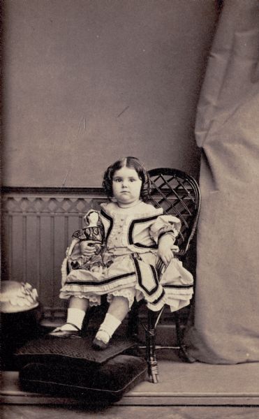 Studio portrait of a young Anita McCormick Blaine (1866-1954) seated in a chair holding a doll. She is wearing a dress and her feet are resting on pillows on the floor.