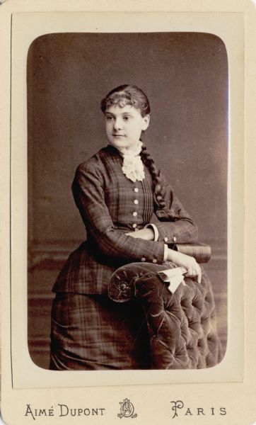 Carte-de-visite studio portrait of Anita McCormick Blaine (1866-1954) as a young adult, posing next to a plush upholstered chair while holding a folded hand fan. She has a bow at the end of her braid, and is wearing a plaid dress with a lace collar.