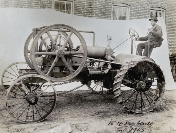 A man is sitting behind the wheel of what appears to be an experimental tractor. The location is staged for a photo shoot, with a white cloth hanging in front of a brick building in the background. The handwritten text on the photograph reads: "15 H.P. Built in 1905."