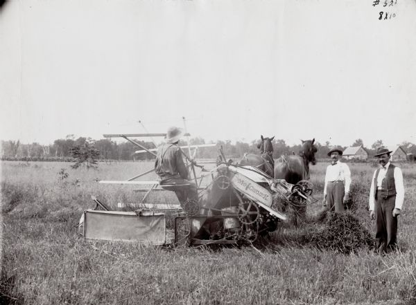A man is operating a McCormick self-binding harvester driven by two horses, while two men are standing beside him in a field. The harvester was built in 1881.
