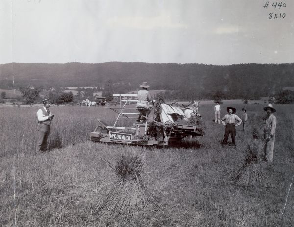 A group of farmers are using a McCormick wire grain binder, built in 1876. A horse-drawn carriage is in the background. The original caption reads: "The improvement embodied in this machine consists of a mechanism for automatically binding the grain with wire, and discharging it in bundles on the ground."