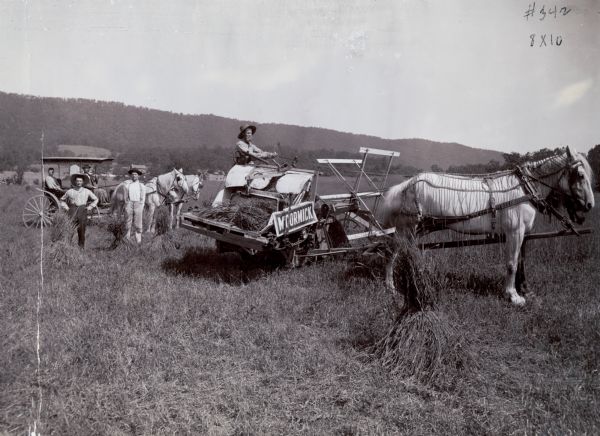 A group of men are using a McCormick wire grain binder led by two horses to harvest a field crop. A horse-drawn carriage is in the background. The original caption reads: "The improvement embodied in this machine consists of a mechanism for automatically binding the grain with wire, and discharging it in bundles on the ground."