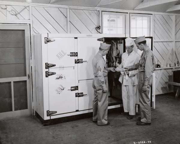 Two uniformed men of the Medical Battalion's Company B stand with the mess sergeant to inspect a piece of poultry from the refrigerator in the mess office of Camp George G. Meade.