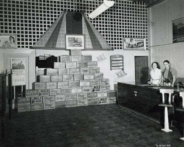52 beverage cases demonstrate the daily capacity of City Coffee Shop's De Luxe cooler. Two women stand behind the counter on the right. Crates include containers for beer, such as Jax, Mule, Stag, Progress, Falstaff and Schlitz.