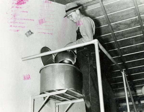 Edgar C. Lyman stands on a platform and uses a stainless steel pail to pour milk into a supply tank.