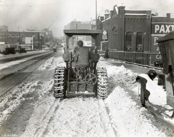Rear view of a man plowing a snow-covered city street with a McCormick-Deering 10-20 industrial tractor. There is a railing on the right.