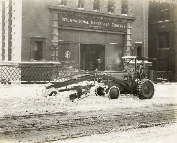A man uses a McCormick-Deering 10-20 industrial tractor to remove snow from the street in front of the International Harvester building.