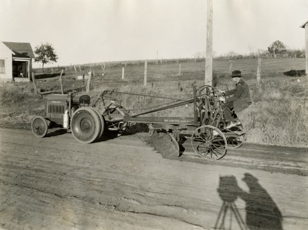 A man uses a McCormick-Deering 10-20 industrial tractor with a grader to smooth a dirt road. Shadows of the photographer and the camera standing on a tripod are in the foreground on the road.