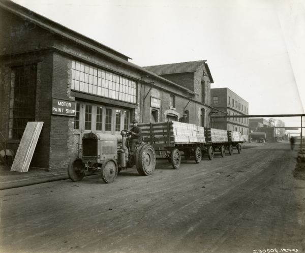 A man is using a McCormick-Deering 10-20 industrial tractor to pull three wagons loaded with boxes. Another man is sitting on top of the boxes. The building on the left has a sign for "Motor Paint Shop."
